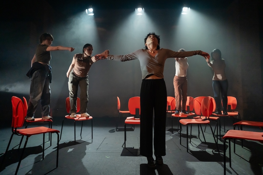 Action shot of the spring 2024 Shakespeare in Performance at RADA final presentation of Julius Caesar. An actor stands with their arms outstretched in the middle of the stage as four actors, standing on bright orange chairs behind them, forcefully point at them.